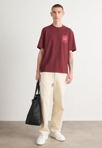 Maroon cotton t-shirt with short sleeves and a logo print, paired with cream-colored trousers and holding a black tote bag. White sneakers included.