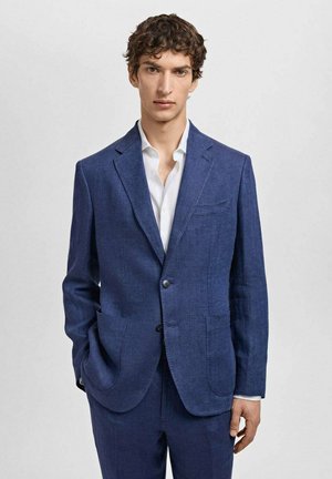 Blazer en lin bleu marine avec revers crantés, deux poches avant et fermeture à un bouton. Porté sur une chemise blanche à col.