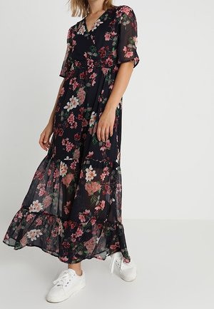 Femme portant une robe maxi noire à fleurs avec manches courtes et baskets blanches, debout devant un fond clair uni.