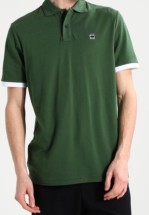 Dunkelgrünes Poloshirt mit kurzen Ärmeln, mit weißen Bündchen und einem kleinen Logo auf der linken Brust. Aus strukturiertem Stoff.
