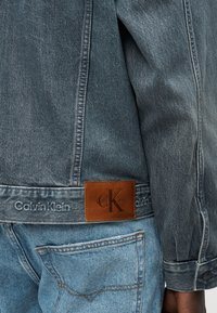 Τζιν μπουφάν με ένθετο λογότυπο Calvin Klein και κεντημένο όνομα της μάρκας, φορεμένο πάνω από ανοιχτό μπλε τζιν τζιν με εμφανή ραφή πίσω τσέπης.