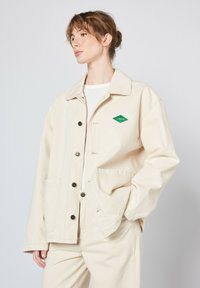 Beige canvasjakke med knaplukning, store påsyede lommer og et grønt diamantlogo. Har en klassisk krave og en afslappet pasform.