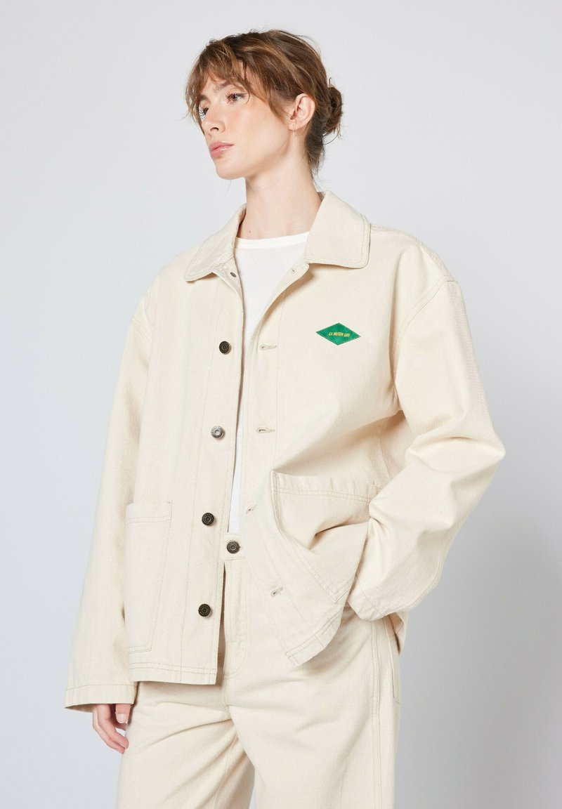 Beige canvasjakke med knaplukning, store påsyede lommer og et grønt diamantlogo. Har en klassisk krave og en afslappet pasform.