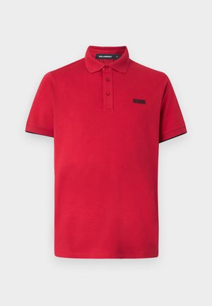 Polo vermelha feita de algodão, com colarinho clássico, dois botões e um pequeno logótipo preto no lado esquerdo do peito. Mangas curtas com acabamento preto.