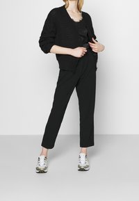 Svart stickad kofta med V-ringning, kombinerad med en svart satintröja, högmidjade svarta byxor och vita sneakers med grå och gula detaljer.