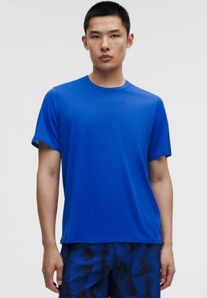 Joven con el pelo negro corto, que lleva una camiseta azul lisa de manga corta y pantalones cortos azul oscuro con estampado, de pie frente a un fondo liso.