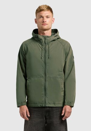 Mand iført olivengrøn hættetrøje med lynlås foran og lommer, stående mod en ensartet lys baggrund.