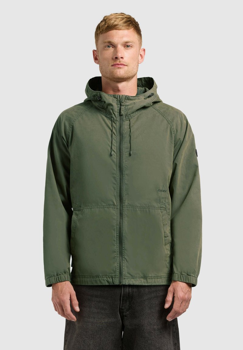 Homme portant une veste à capuche vert olive avec fermeture éclair frontale et poches, debout devant un fond clair uni.