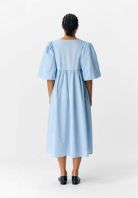 Robe midi bleu clair avec des manches bouffantes, taille froncée et texture lisse. Encolure ronde simple et silhouette fluide. Chaussures noires.