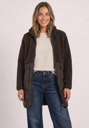Femme souriante aux cheveux mi-longs porte une longue veste polaire marron foncé sur un haut beige et un jean bleu, debout devant un fond uni.