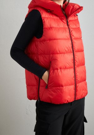 Persoon draagt een felrode, gewatteerde mouwloze jas over een zwart shirt met lange mouwen, met één hand in de zak van de jas.