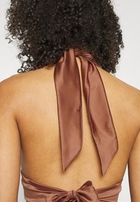 Top halter in satin marrone con lunghe cravatte sul retro, caratterizzato da una texture liscia e un design con schiena scoperta, che mette in mostra la pelle e i capelli.