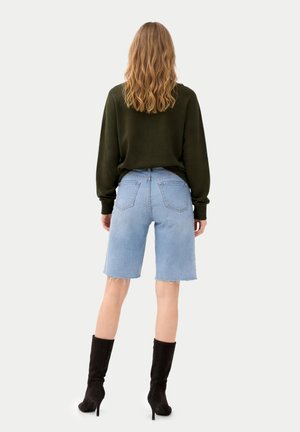 Frau mit welligem Haar, die einen dunkelgrünen Pullover, kniehohe hellblaue Jeansshorts mit rohen Säumen und schwarze Stiefel mit Absatz trägt, steht mit dem Rücken zu uns.