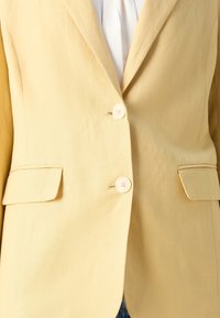 Blazer jaune clair avec deux boutons et poches à rabat, porté sur un haut blanc.