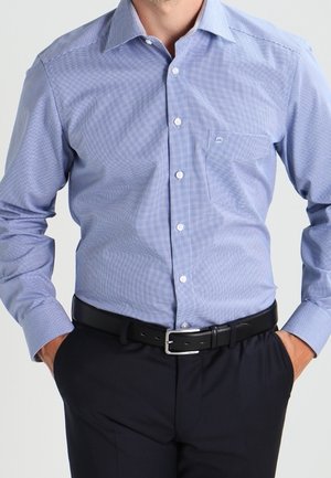 Mand iført en lyseblå ternet skjorte med lange ærmer, knappet op, stukket ned i mørke bukser med sort bælte, hænderne i lommerne.