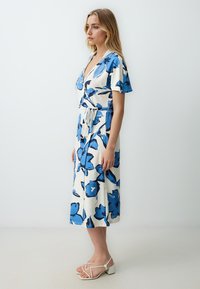 Bloemige wikkeljurk met korte mouwen, met blauwe en witte bloemenpatronen, een strik in de taille en een jurk tot op de knie. G gedragen met sandalen met banden.