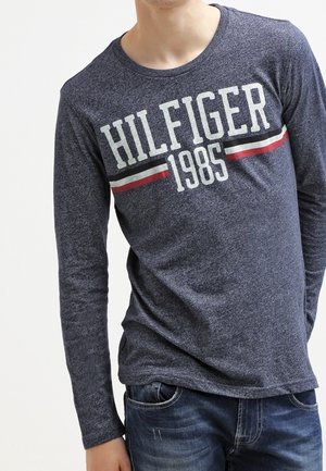 Mand iført en marineblå langærmet skjorte med "HILFIGER 1985" og røde, hvide og sorte striber på tværs af brystet, kombineret med blå jeans.