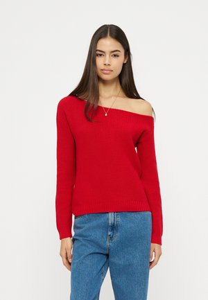 Žena s dlouhými tmavými vlasy, která má na sobě červený svetr s odhalenými rameny a modré džíny, stojící před jednoduchým bílým pozadím.