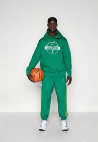 Žalias Nike džemperis su „Celtics“ logotipu, derančios žalios kelnės su šviesiai žaliomis šoninėmis juostelėmis ir balti sportbačiai, laiko krepšinio kamuolį.