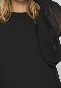 Top nero con un corpo in cotone solido e maniche trasparenti e puffate. Il collo è arrotondato e le maniche sono arricciate ai polsini.