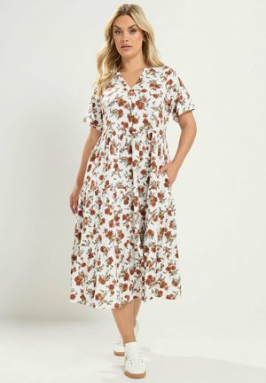 Vrouw met lang blond haar die een witte midi-jurk met bloemenprint en korte mouwen en witte sneakers draagt, staand met één hand in haar zak.