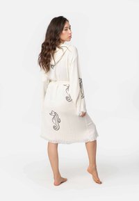 Biela kapucňová róba s čiernym vyšívaným morským koníkom, strapcovým lemom a viazaním. Mäkká, textúrovaná tkanina s uvoľneným strihom, dĺžka po členky.