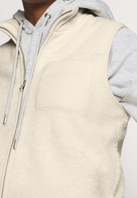 Beżowa kamizelka polarowa z zamkiem z przodu, małą kieszonką na piersi i miękką fakturą, noszona na szarej bluzie z kapturem.