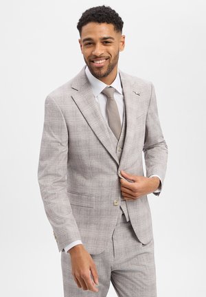 Mann lächelt, richtet den Knopf an einem hellgrauen karierten Anzug mit passender Weste und Krawatte, steht vor einfachem Hintergrund.