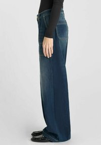 Jean en denim bleu foncé à jambes larges avec poches avant et arrière et coutures visibles. Porté avec des ballerines noires, affiché de côté.