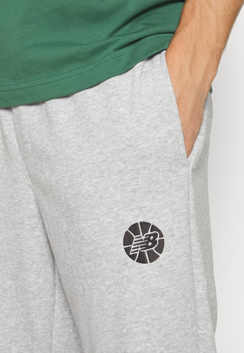 Graue Jogginghosen mit einer weichen Textur verfügen über ein schwarzes New Balance-Logo am linken Oberschenkel und seitliche Taschen. Kombiniert mit einem grünen Shirt.