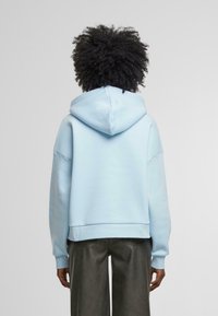 Ljusblå, croppad hoodie gjord av mjukt tyg, med stor huva, sänkta axlar och ribbade ärmslut. Matt yta och minimala design detaljer.