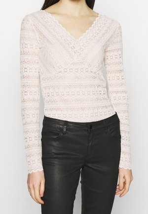 Femme portant un haut blanc en dentelle à manches longues avec un col en V, accompagné d'un pantalon en cuir noir ajusté, debout devant un fond uni.