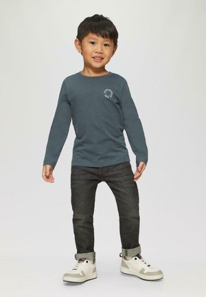 Ein kleiner Junge steht da und trägt ein dunkelblaues Langarmshirt, hochgekrempelte dunkle Jeans und weiße Sneakers vor einem einfachen hellen Hintergrund.