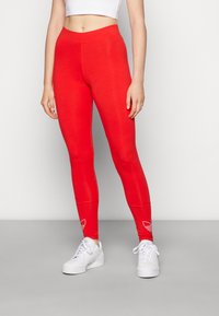 Rode leggings van rekbare stof met een hoge taille, gecombineerd met witte sneakers. Beschikt over een eenvoudig logo bij de enkels.