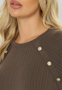 Blond kvinna med en ribbad brun topp med dekorativa guldknappar längs axelsömmen, läpparna lätt öppna.