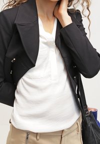 Blazer negro recortado con solapas y detalles metálicos plateados, superpuesto sobre una camisa blanca. Pantalones beige visibles, con un bolso negro al fondo.