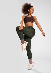 Sujetador deportivo marrón y leggings verdes oliva con acentos texturizados. Zapatillas blancas con detalles morados. Modelo posa de manera dinámica.
