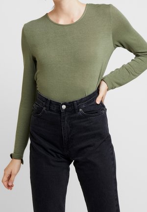 Osoba w oliwkowozielonej koszuli z długim rękawem, wpuszczonej w wysokowysokie czarne jeansy, stojąca z jedną ręką na biodrze, drugą swobodnie opadającą.