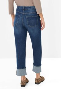 Frau in dunkelblauen, umgekrempelten Jeans mit Gesäßtaschen und Loafern im Leopardenmuster, die vor einem weißen Hintergrund steht.