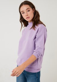 Felpa lilla con colletto alto, polsini e orlo a coste, realizzata in tessuto morbido. Il modello la indossa sopra jeans blu.