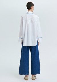 Camisa blanca de botones con Acabados azules y botones dorados, combinada con pantalones de mezclilla oscura de pierna ancha y calzado beige. Textura suave.