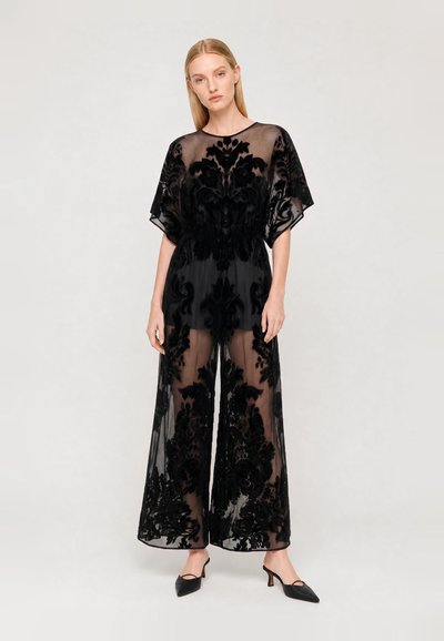 Blonde woman stands wearing a black sheer jumpsuit with floral velvet patterns and black low-heeled pointed shoes against a plain background.