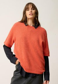 Suéter de manga corta en color coral naranja, hecho de un material suave y peludo, con un dobladillo redondeado, usado sobre una camisa negra con cuello y unos jeans grises.