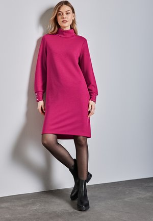 Fuchsia polotröja klänning med långa ärmar, ribbad textur och knappdekorationer vid ärmsluten, tillsammans med svarta ankelstövlar och genomskinliga strumpbyxor.