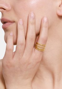 Bague en métal doré avec plusieurs bandes empilées, présentant des surfaces alternées lisses et texturées. Gros plan sur le ton de la peau, mettant en valeur les détails de la main.