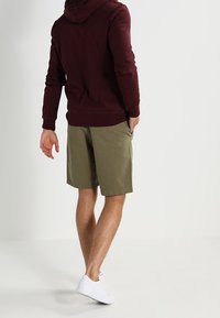 Persona in piedi con le spalle rivolte all'osservatore, che indossa un hoodie bordeaux, pantaloni corti verdi oliva e sneaker bianche, su uno sfondo neutro.