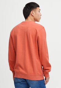 Sudadera naranja con cuello redondo y mangas largas. Textura suave, corte regular y puños acanalados. Llevada sobre unos jeans azules, vista desde la parte posterior.