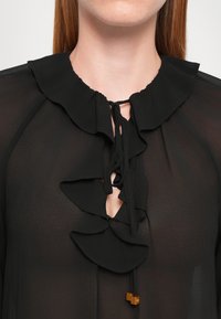 Svart bluse med rynkede detaljer ved halsen og en knytedetalj i midten. Lett stoff med en jevn tekstur og subtil glans.