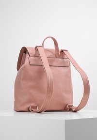 Mjukt rosa läderryggsäck med justerbara remmar och ett handtag på toppen, stående på en vit sockel mot en grå bakgrund.