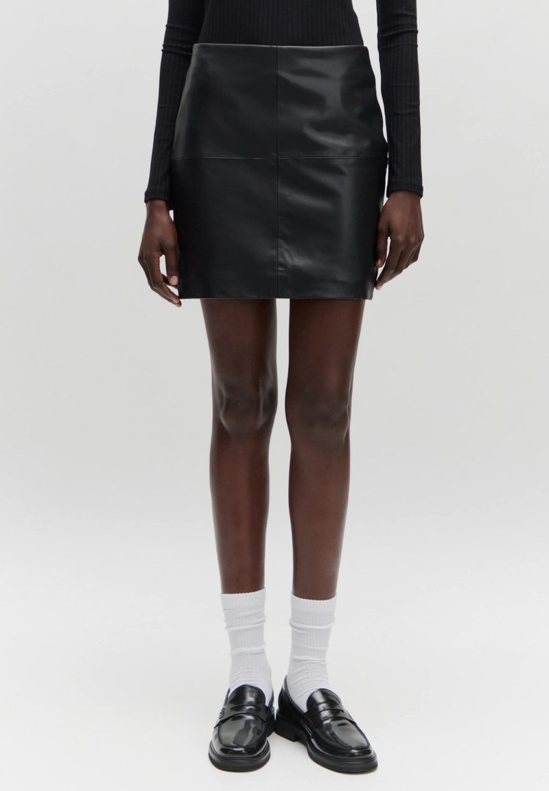 Femme portant une mini-jupe en cuir noire, un haut noir à manches longues, des chaussettes blanches et des mocassins en cuir noirs, debout sur un fond uni.